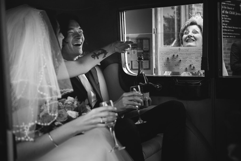 Black and white: a bride and groom laughing in a wedding car as a guest leans in at the window