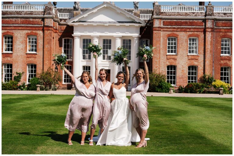 A bride and three bridesmaids raising their bouquets on the lawn in front of a red brick country house