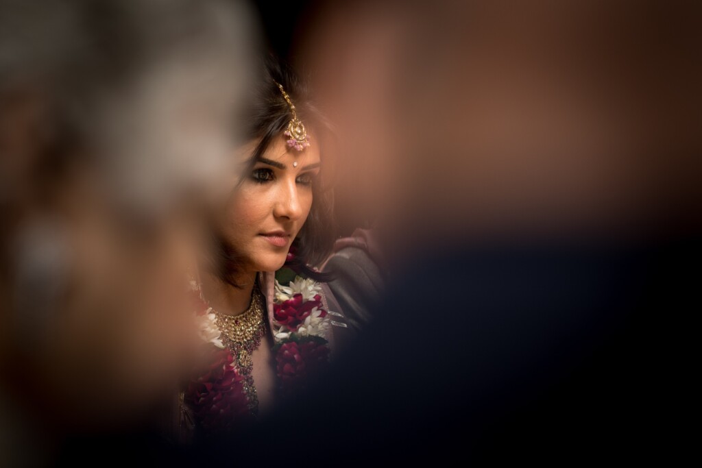 A bride in a gold forehead ornament seen through a gap between blurred guests