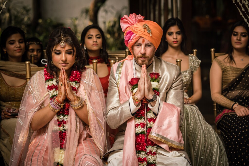 A bride and groom in rose garlands sitting with their palms pressed together as women seated behind them look on