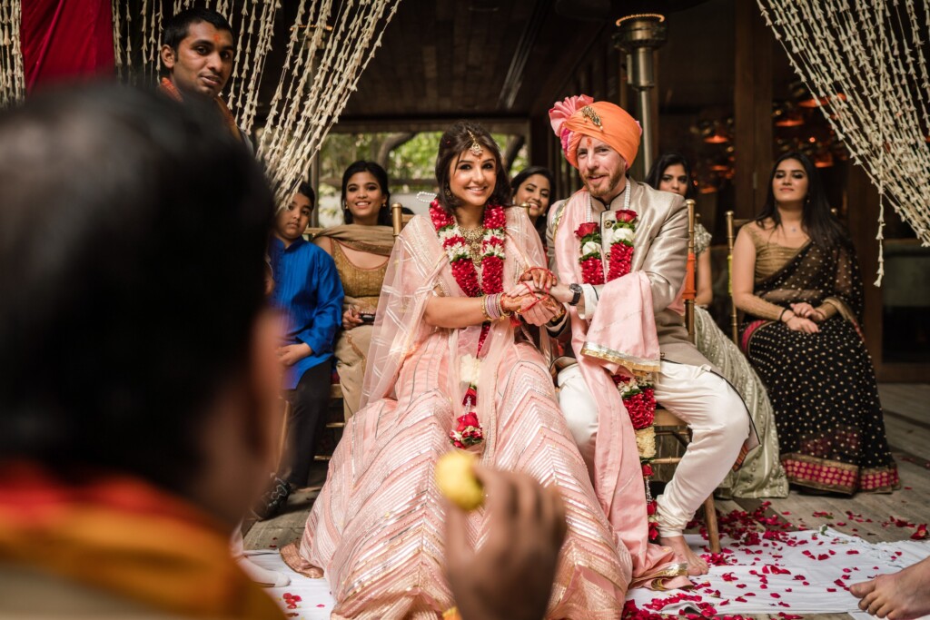 A bride and groom in garlands seated hand in hand during the ceremony as guests watch, a blurred guest in the foreground