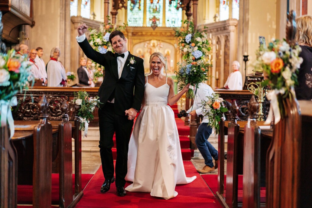A groom punching the air as he walks back down the aisle hand in hand with his laughing bride