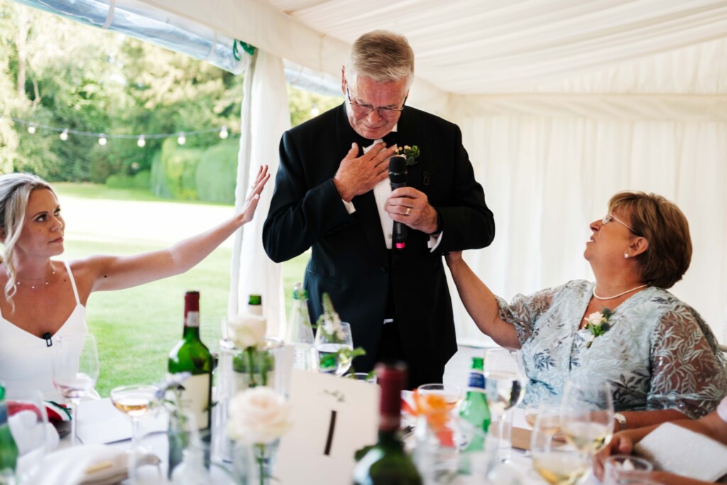 A man with his hand on his chest during his speech in a marquee as the bride and an older woman reach out to him