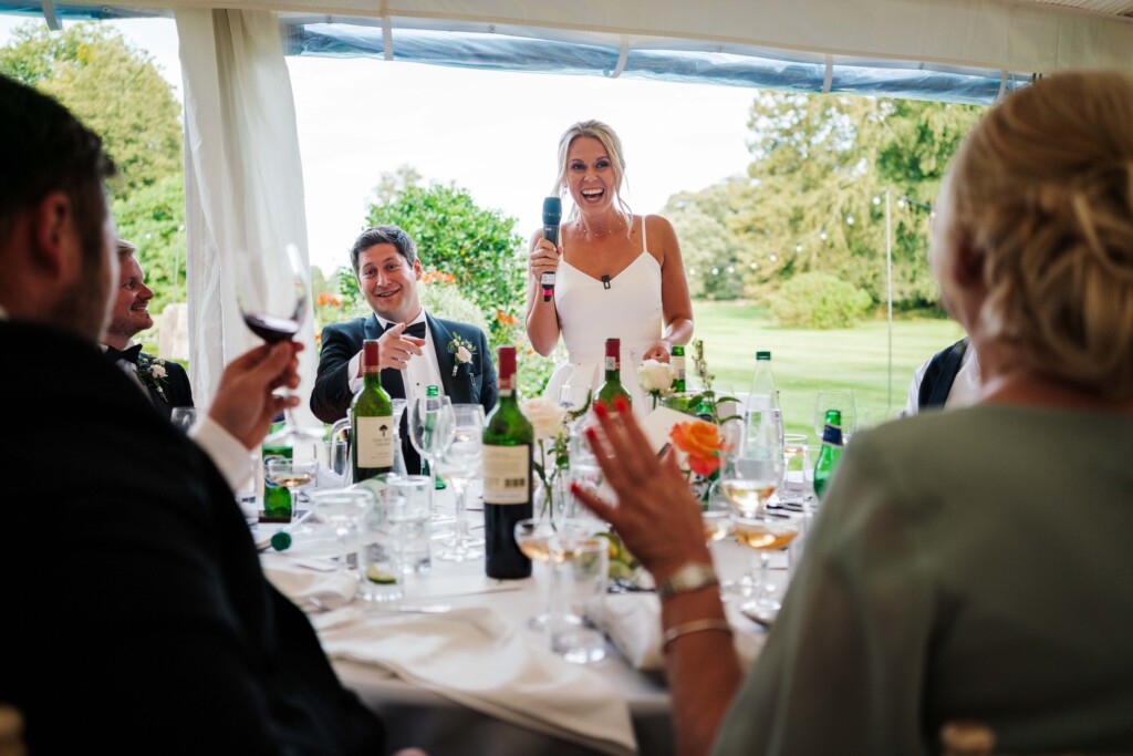 A laughing bride giving a speech in a marquee as the groom and guests laugh at their table