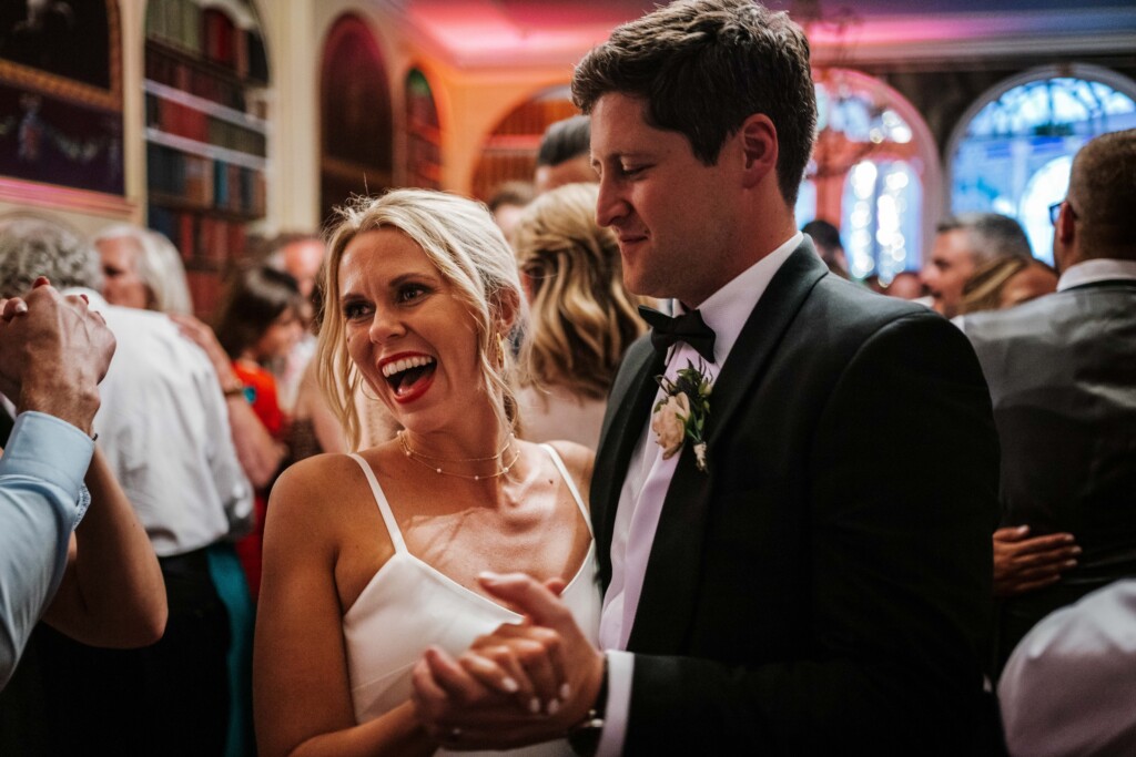 A laughing bride and groom clapping among guests on a crowded dance floor in a library
